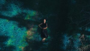 Aerial view of a person lying on their back in a dark, grassy clearing, wearing a black outfit, surrounded by blue-green vegetation.