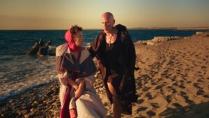 Two costumed people walking along a rocky beach at sunset, one in a pink hooded cloak and the other in a black cape with jewelry on the chest area.