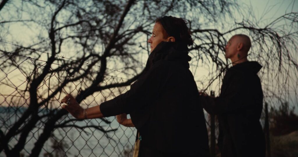 Two people in dark clothing stand by a chain-link fence topped with barbed wire at dusk.