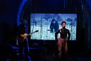 Singer at a microphone with hands on hips on a dim stage; guitarist in blue light to the left, with a forest-scene projection behind them