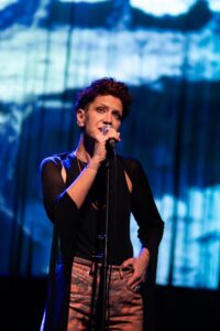 Female singer on stage, mic in hand, wearing a black top with sheer sleeves and patterned pants, blue abstract backdrop.