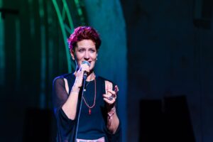 Female singer on stage holds microphone and sings, illuminated by blue and green stage lights.