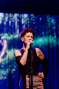 Female singer performing on stage, gripping a microphone with eyes closed under blue-purple lights.